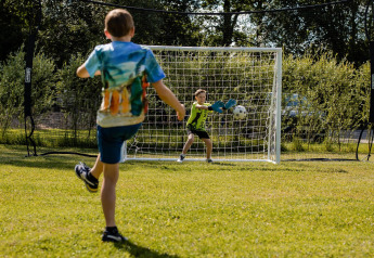 Twee kinderen spelen voetbal bij een doel op het gras bij een safaritent, omgeven door bomen.