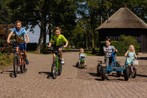 Kinderen fietsen en rijden op skelters bij een safaritent op een zonnige, groene camping.