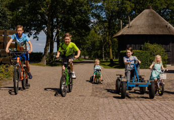 Kinderen fietsen en rijden op skelters bij een safaritent op een zonnige, groene camping.