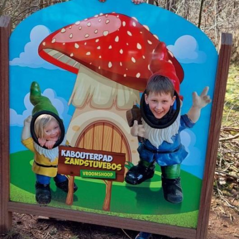 Niños posan sonrientes con caras en un cartel de duendes en Kabouterpad Zandstuvebos, Vroomshoop.