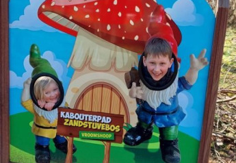 Niños posan sonrientes con caras en un cartel de duendes en Kabouterpad Zandstuvebos, Vroomshoop.