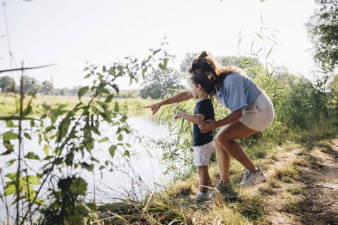 Een vrouw en een kind staan aan de rand van een meer in de natuur, de vrouw wijst iets aan op het water.