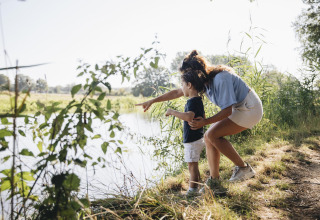 Een vrouw en een kind staan aan de rand van een meer in de natuur, de vrouw wijst iets aan op het water.