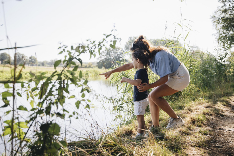 A woman and a child stand by a lakeside in nature, the woman pointing across the water in the distance.