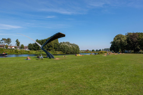 Campo verde junto a un río con personas descansando y una escultura moderna en un glamping.