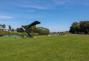 Campo verde junto a un río con personas descansando y una escultura moderna en un glamping.