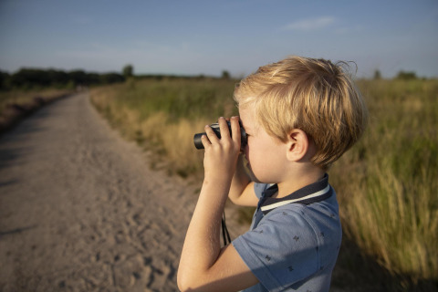 Jongen met verrekijker op safaritent-uitje, kijkend over een zandpad in een groen natuurgebied.