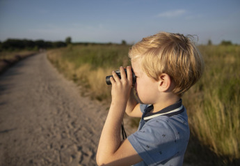 Jongen met verrekijker op safaritent-uitje, kijkend over een zandpad in een groen natuurgebied.