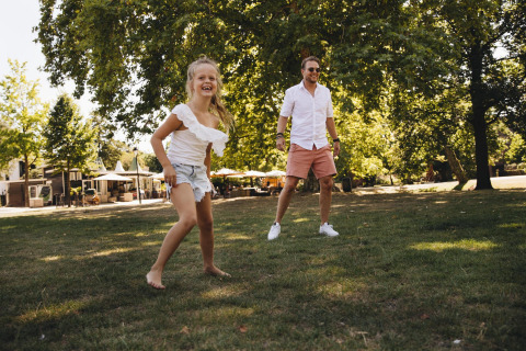 Un homme souriant et une fille jouent pieds nus sur l’herbe devant des tentes safari dans un parc ensoleillé.