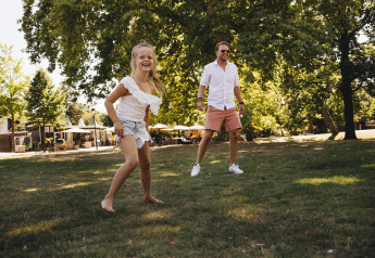 Un homme souriant et une fille jouent pieds nus sur l’herbe devant des tentes safari dans un parc ensoleillé.
