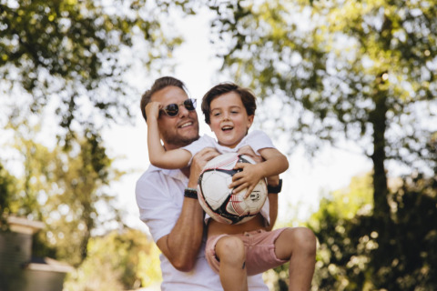 Een lachende man tilt een blij kind met een voetbal buiten onder de bomen op een zonnige dag.