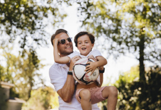 A smiling man lifts a happy child holding a soccer ball outdoors under trees on a sunny day.