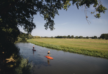 Two people paddleboarding on a calm river beside green fields and trees, near a safari tent location.