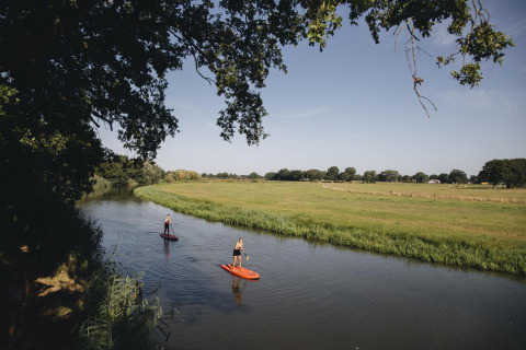 Two people paddleboarding on a calm river beside green fields and trees, near a safari tent location.