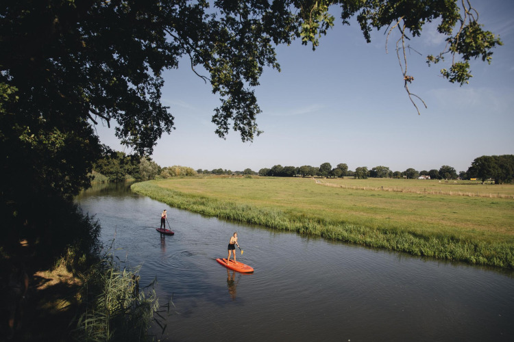 Zwei Menschen paddeln auf Boards einen ruhigen Fluss entlang, umgeben von Wiesen nahe einem Safarizelt.