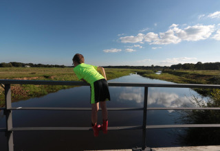 Jongen met felgroene outfit leunt over de brugleuning aan een rivier bij een safaritentlocatie.