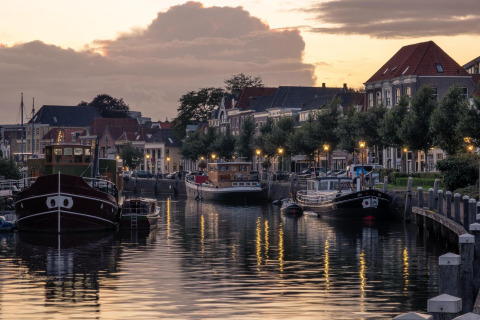 Avondbeeld van glamping accommodatie aan een kanaal met boten, lantaarns en Hollandse huizen.