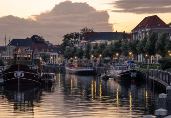 Avondbeeld van glamping accommodatie aan een kanaal met boten, lantaarns en Hollandse huizen.