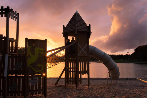 Speeltuin bij glampingaccommodatie aan het meer bij zonsondergang, kinderen spelen op toren en glijbaan.