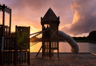 Zona de juegos en alojamiento glamping junto al lago al atardecer, con niños jugando en la torre y tobogán.
