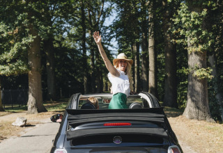 Vrolijke vrouw met hoed staat in een cabriolet tussen de bomen, genietend van een glampingvakantie.