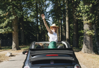 Vrolijke vrouw met hoed staat in een cabriolet tussen de bomen, genietend van een glampingvakantie.