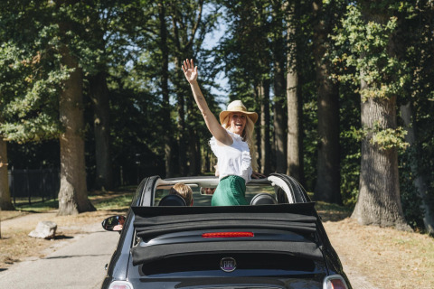 Happy woman in a sunhat stands in a convertible, waving and smiling during a glamping stay.