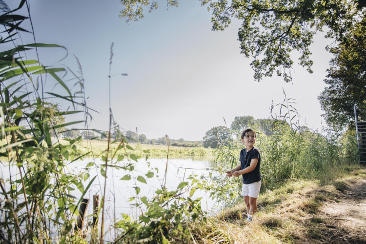 Child fishing by a river near a safari tent at CharmeCamping De Regge-Vallei in the Netherlands, sunny day.