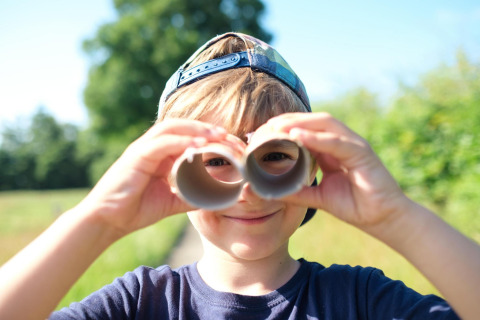 Boy in cap playing outdoors at a safari tent, pretending to use binoculars made from cardboard tubes