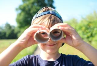 Jongen met petje bij safaritent, kijkt door kartonnen buisjes als verrekijker, zomerse buitenscène