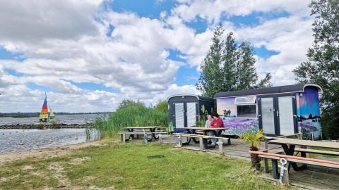 Two people sit at a picnic table by the Tiny Wagon at Marina Parcs Almere, Netherlands, near a lake.