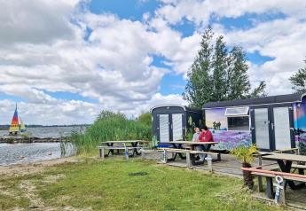 Two people sit at a picnic table by the Tiny Wagon at Marina Parcs Almere, Netherlands, near a lake.