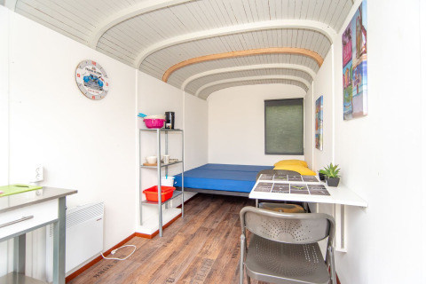 Vista interior del Tiny Wagon en Marina Parcs Almere, Países Bajos, con cama, escritorio y estanterías.