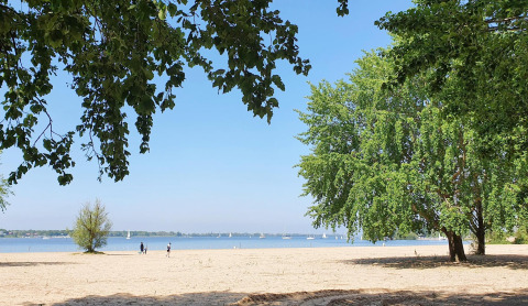 Zandstrand met groene bomen en wandelaars, uitkijkend op een meer met zeilboten in de verte.