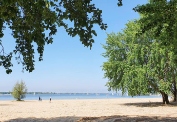Strandudsigt med sand, grønne træer og folk der går, udsigt til vandet og sejlskibe i det fjerne.