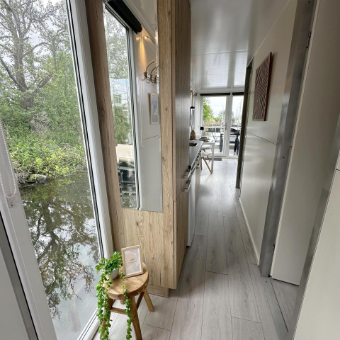 Indvendig gang i husbåd med store vinduer ved vandet, lyst træinteriør og udsigt til natur, Almere.