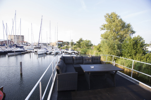 Terraza de casa flotante con sofá y mesa, junto a un puerto deportivo con veleros en Almere, Países Bajos.