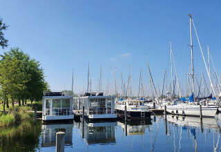 Woonboot aan Marina Parcs Almere, Nederland, omringd door zeilboten en spiegelend water onder blauwe lucht.