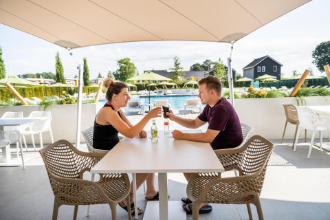 To mennesker skåler ved et udendørs bord på Resort Mooi Bemelen, med pool og grøn udsigt i baggrunden.
