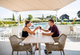 Dos personas brindan en una mesa al aire libre en Resort Mooi Bemelen, con piscina y jardín de fondo.