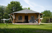 Tienda safari con porche cubierto, barbacoa, muebles de exterior y hamaca en césped rodeado de naturaleza.