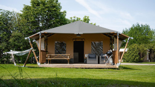 Tenda safari con veranda coperta, barbecue, poltrone da esterno e amaca su prato verde in mezzo alla natura.