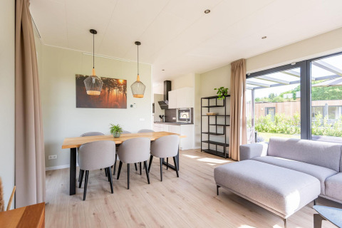 Moderne lodge med åbent opholdsrum, spisebord, sofa og store vinduer med udsigt til haven om dagen.