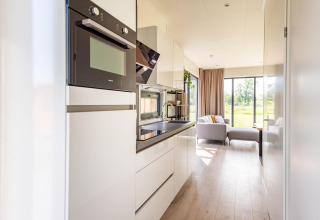 Moderne lodge-keuken met witte kasten, ingebouwde toestellen en zicht op een lichte woonkamer en tuin.
