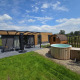 Buitenzicht op een moderne lodge met terras, houten sauna en jacuzzi op een groen gazon.