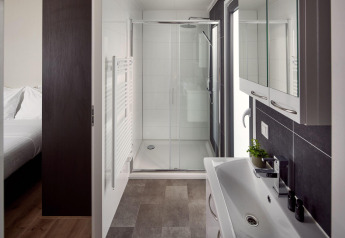 Moderne badeværelse på lodge med glasbruser, stort spejl, vask, mørke fliser og tilstødende soveværelse.