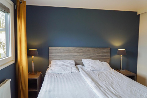 Dormitorio moderno en una cabaña Comfort de EuroParcs Noordwijkse Duinen con cama doble, lámparas y pared azul.