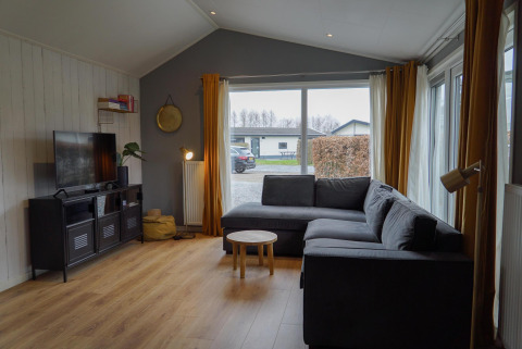 Gemütliches Wohnzimmer einer Hütte im EuroParcs Noordwijkse Duinen mit grauem Sofa und großen Fenstern.