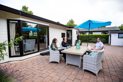 Quatre personnes sont assises dehors autour d’une table devant un chalet à EuroParcs Noordwijkse Duinen.