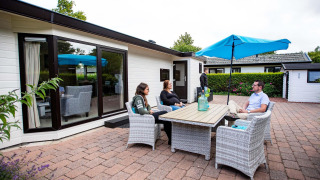 Quatre personnes sont assises dehors autour d’une table devant un chalet à EuroParcs Noordwijkse Duinen.